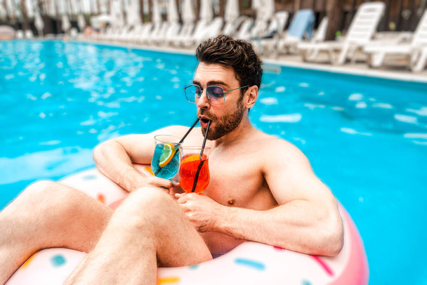 Handsome bearded guy drinking two cocktails while spending his holiday in the swimming pool. Summertime, holidays, lifestyle concept - Photo, Image