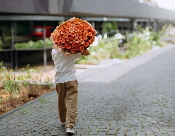 Ein kleiner Junge trägt seiner Freundin einen großen Strauß Rosen. Junge mit Rosenblüten, Rücken. geht mit Blumen hinterher - Foto, Bild
