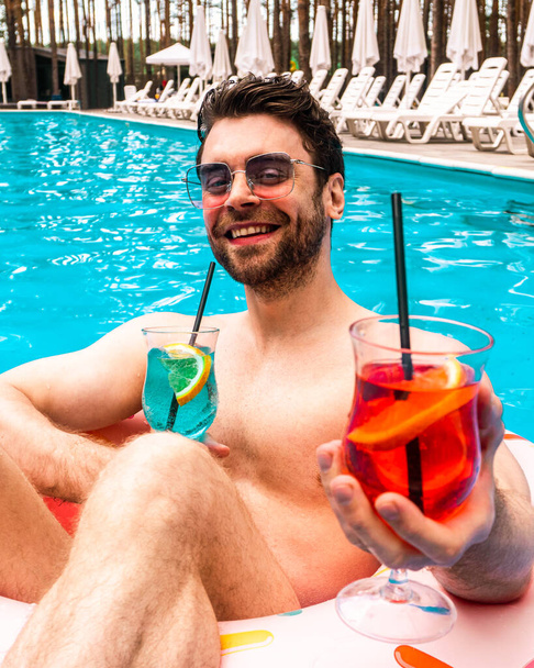 Χαρούμενος όμορφος τύπος που απολαμβάνει το Σαββατοκύριακο στην πισίνα έξω ενώ κρατάει δύο κοκτέιλ, ηλιόλουστη μέρα. Καλοκαίρι, διακοπές, τρόπος ζωής έννοια - Φωτογραφία, εικόνα