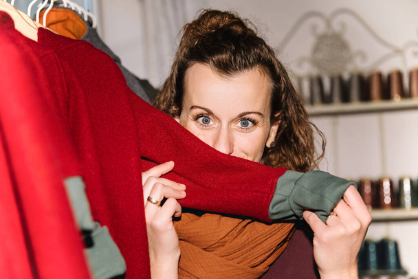Eine fröhliche Frau mit lockigem Haar versteckt sich spielerisch hinter einem roten Kleidungsstück und zeigt ihre Augen in einem Boutique-Ambiente vor gemütlicher Kulisse - Foto, Bild