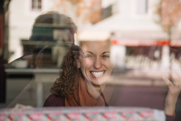 Mujer joven radiante sonriendo ampliamente detrás de una ventana reflectante, café urbano borrosa en el fondo con tonos otoñales - Foto, imagen