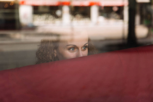 Donna riflessiva con i capelli ricci guardando fuori da dietro una finestra di vetro riflettente, la vita di città visibile sullo sfondo soft focus - Foto, immagini