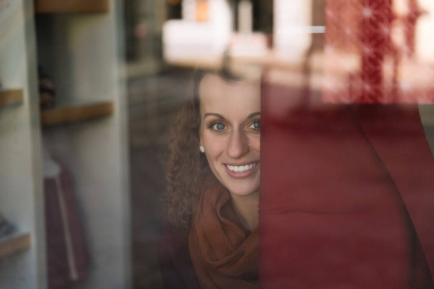 Bezauberndes Lächeln einer jungen Frau mit lockigem Haar durch ein Café-Fenster gesehen, warmes Tageslicht erhellt ihr Gesicht - Foto, Bild