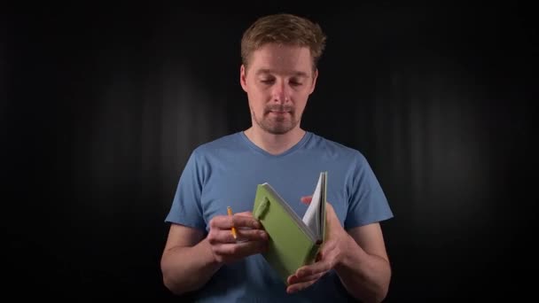 A portrait of a handsome young white man holding a notebook, opening it, jotting down notes, making entries in a planner on a black background. Planning and record-keeping in a purposeful lifestyle - Footage, Video