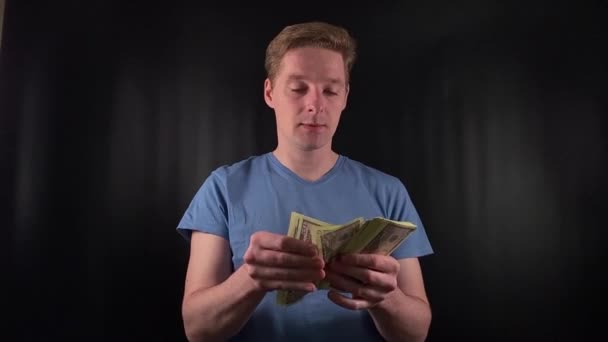Czarujący młody człowiek z entuzjazmem liczy stos banknotów dolarowych. Dreszcz szczęścia na prostym, czarnym tle. Uosobienie radości uroczystości po trafieniu w dziesiątkę - Materiał filmowy, wideo