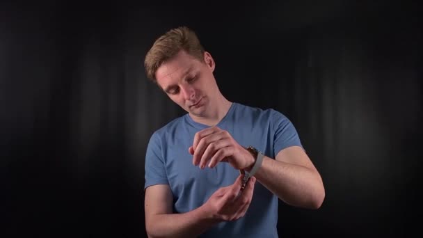 a stylish young man deliberately fastens an elegant timepiece around his wrist on the minimal black backdrop. His purposeful movements exude an unmistakable air of poise and man-of-the-world flair - Footage, Video