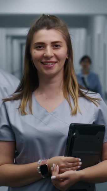 Ženský doktor stojí v moderní chodbě kliniky. Profesionální zdravotník drží digitální tablet, usmívá se a dívá se do kamery. Lékařský personál a pacienti v zázemí v nemocniční nebo lékařské centrum chodby. - Záběry, video