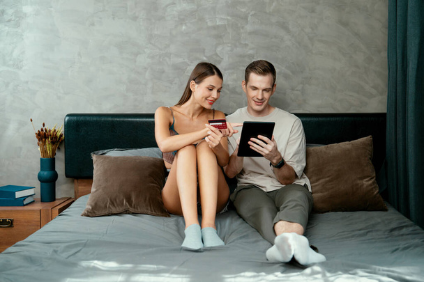 Nuori pari istua kotona makuuhuoneessa käyttäen online-maksu sovellus ja digitaalinen lompakko älypuhelin maksaa luottokortilla. Sähköinen kaupankäynti ostoksia ja moderni hankinta mobiili internet. Paljastaminen - Valokuva, kuva