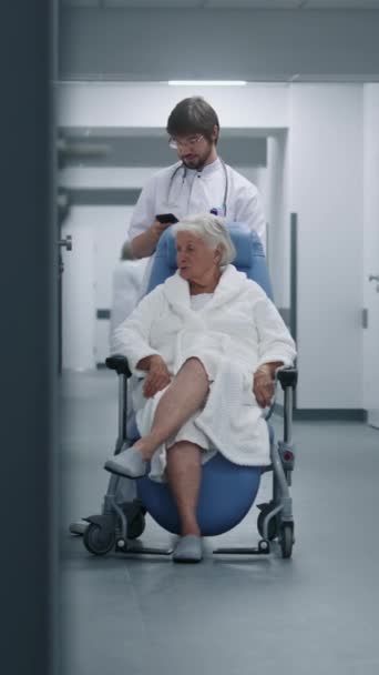 Il medico maschio sta con la donna anziana in sedia a rotelle nel corridoio della clinica, usa il telefono cellulare. Medic con paziente femminile vicino sala operatoria o procedure. Personale medico e pazienti nel corridoio dell'ospedale. - Filmati, video