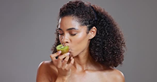 Gesicht, Schönheit und Frau essen Kiwi im Studio auf grauem Hintergrund für Gesundheit, Vitamin C oder Wellness. Porträt, Frucht für die Hautpflege und glückliches junges Model mit natürlicher Nahrung zur Entgiftung, Diät oder Ernährung. - Filmmaterial, Video