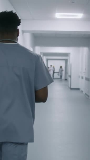 Un spécialiste afro-américain marche dans le couloir de l'hôpital. Homme médecin va sur rendez-vous avec les patients. Personnel médical multiethnique au travail dans une clinique moderne. Tir vertical. Vue arrière. - Séquence, vidéo