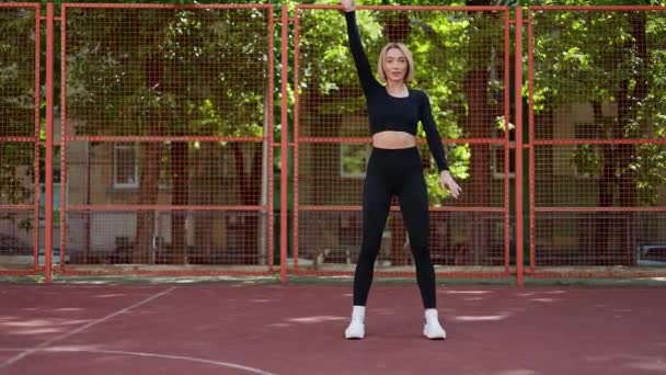 Uma jovem mulher em traje fitness estende os braços em uma quadra de basquete, se preparando para seu treino. - Filmagem, Vídeo
