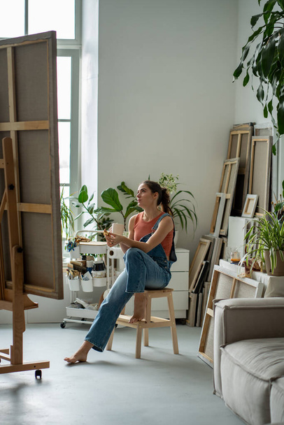 Przemyślana artystyczna kobieta robi sobie przerwę od rysowania, odpoczywając na krześle, podjadając bułeczki, pijąc herbatę, kawę. Poważna zamyślona kobieta patrzy na malowanie podczas lunchu, planując kolejny etap pracy - Zdjęcie, obraz