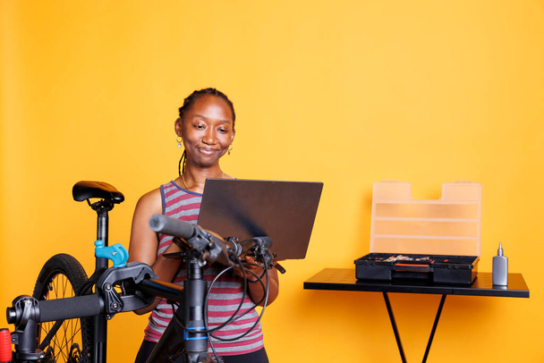 Przed żółtym tle, czarna kobieta wykonuje konserwację roweru przy użyciu laptopa cyfrowego dla instrukcji. Afroamerykanka korzystająca z minikomputera i profesjonalnych narzędzi do mocowania rowerów. - Zdjęcie, obraz