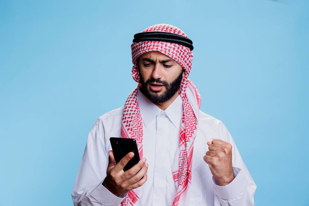 Podekscytowany muzułmanin noszący islamskie tradycyjne ubrania czytający wiadomości w mediach społecznościowych za pomocą smartfona. Arab ubrany w szalik i thobe ghutry podczas sprawdzania wiadomości przez telefon komórkowy - Zdjęcie, obraz