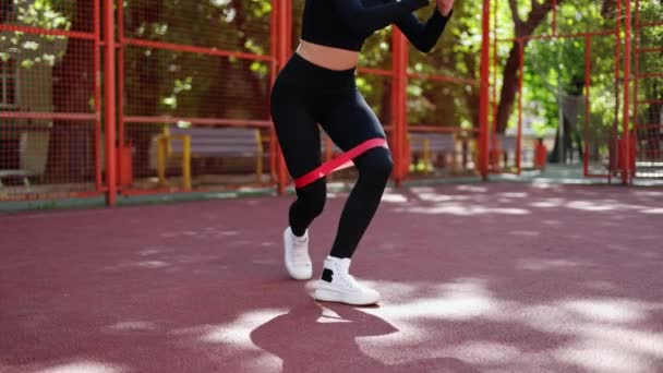 Vrouw in zwarte sportkleding doet beenoefeningen met een rode weerstandsband op een sportveld. - Video