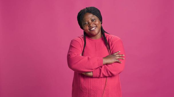 Enthousiaste zwarte vrouw staat voor een levendige roze achtergrond met armen gekruist op haar borst. Afro-Amerikaanse fashionista elegant poseren tegen geïsoleerde roze achtergrond. - Foto, afbeelding