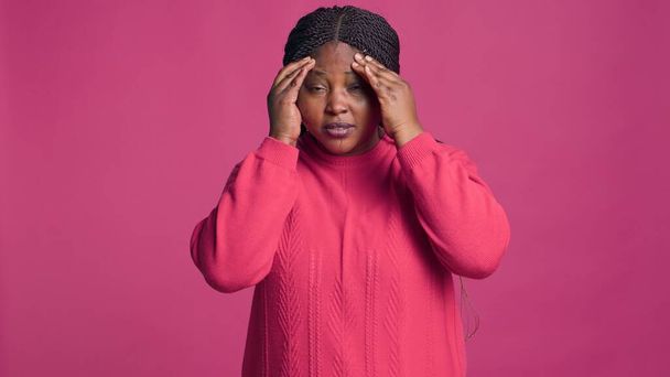 Mulher negra na moda vestindo suéter rosa sofrendo uma dor de cabeça ruim. Feminino Africano americano beleza cobrindo rosto na frente da câmera mostrando ter uma enxaqueca. - Foto, Imagem