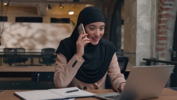 Muzułmańska arabska islamska kobieta w hidżabie szczęśliwa businesswoman rozmawiać telefon komórkowy praca w biurze firmy kobieta konsultant menedżer biznes dziewczyna konsultować klienta online za pomocą laptopa napisać zadanie uśmiech - Materiał filmowy, wideo