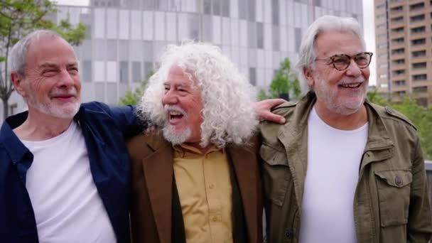 Group of three mature caucasian men enjoying embracing walk together laughing outdoor. Senior male old friends having fun hugging strolling happy in the urban city street. Elderly people golden ager - Footage, Video