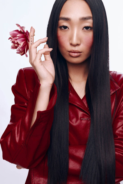 Tajemnicza kobieta z długimi czarnymi włosami i czerwoną skórzaną kurtką trzymającą kwiat przed twarzą - Zdjęcie, obraz