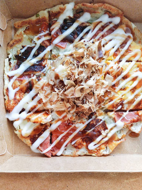 Typischer japanischer Straßensnack, Okonomiyaki in einer Verpackungsbox. Okonomiyaki wird aus Weizenmehl hergestellt, das Gemüse, Fleisch, Fisch usw. enthält. Dann auf einer Eisenplatte gebacken und mit Sauce beschmiert - Foto, Bild