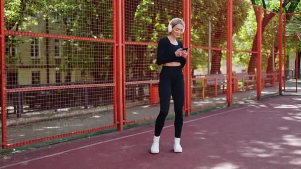 Sorrindo mulher esportiva em sportswear ouve música em fones de ouvido enquanto usa seu smartphone em um campo de basquete - Filmagem, Vídeo