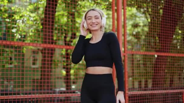 Die fitte junge Frau im schwarzen Trainingsoutfit genießt die Musik auf ihren Kopfhörern und tanzt draußen vor einem roten Zaun.. - Filmmaterial, Video