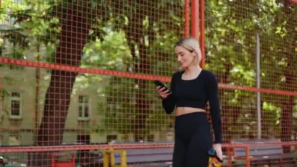 Молодая женщина в спортивной одежде прогуливается по забору на спортивной площадке, проверяет телефон и улыбается после тренировки. - Кадры, видео