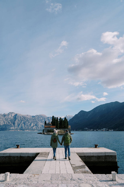 Άντρας και γυναίκα περπατούν κρατώντας τα χέρια κατά μήκος της προβλήτας με θέα το νησί του Αγίου Γεωργίου. Πίσω όψη. Μαυροβούνιο. Υψηλής ποιότητας φωτογραφία - Φωτογραφία, εικόνα