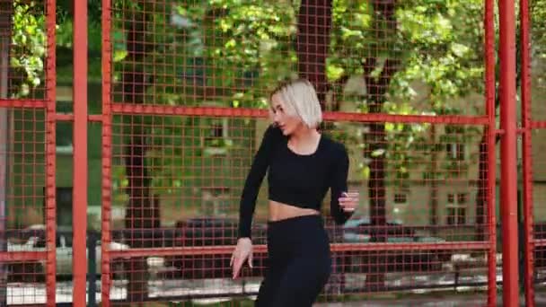Genç bir kadın ısınıyor ve parkta basketbol sahasının yanında geriniyor. - Video, Çekim