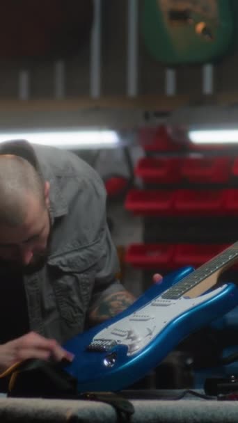 Επαγγελματική αρσενικό ξυλουργός σκουπίζει χορδές κιθάρα με κουρέλι και λεπτομερείς επιθεωρήσεις κιθάρα. Ξυλουργός τεχνίτης, σχεδιαστής κάνει ξύλινα μουσικά όργανα στο σύγχρονο εργαστήριο. Κάθετη βολή - Πλάνα, βίντεο