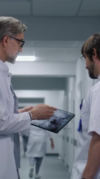 El médico varón muestra una resonancia magnética, una imagen de tomografía computarizada del cerebro al médico colega usando una tableta digital. Hablan los médicos, se paran en el pasillo de la clínica moderna. Personal médico y pacientes en el pasillo del hospital o centro médico. - Metraje, vídeo