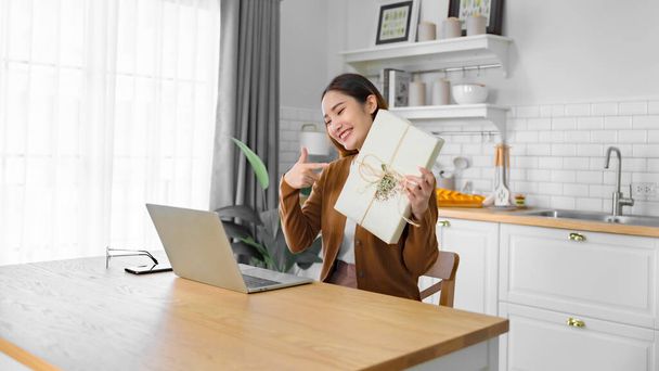 Молода азіатка використовує комп'ютерний ноутбук вдома. Жінка показує подарункову коробку під час відео розмови з другом. З Днем Народження, З Новим роком, З Днем Подяки
 - Фото, зображення