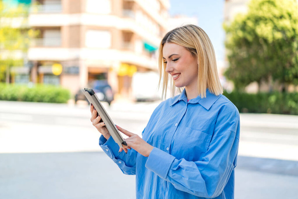 Jeune jolie femme blonde à l'extérieur touchant l'écran de la tablette avec une expression heureuse - Photo, image