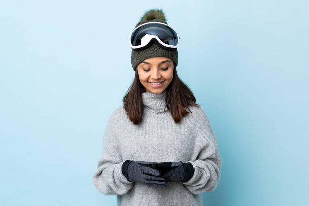 Menina esquiador de corrida mista com óculos de snowboard sobre fundo azul isolado enviando uma mensagem com o celular. - Foto, Imagem