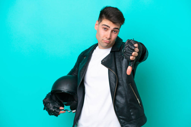 Joven hombre caucásico con casco de moto aislado sobre fondo azul mostrando el pulgar hacia abajo con expresión negativa - Foto, Imagen