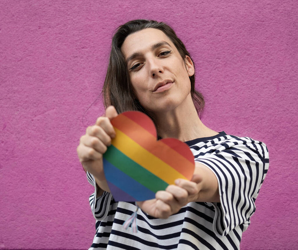 Vista frontal cercana de una mujer lesbiana con un corazón de arco iris para apoyar a la comunidad LGBT aislada sobre un fondo rosa. - Foto, imagen
