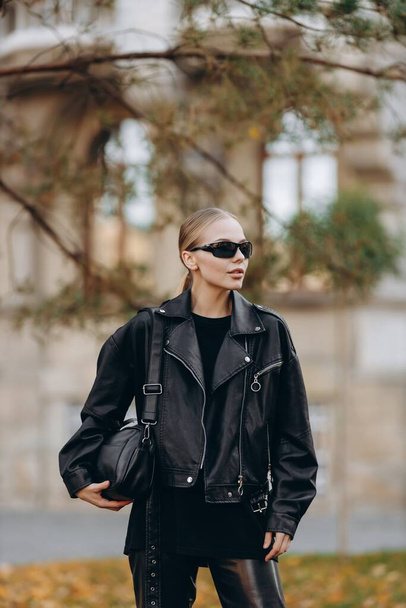 Femme à la mode avec des lunettes de soleil noires sur les yeux regarde loin de la caméra et porte un t-shirt de base noir, veste en cuir noir et pantalon en cuir noir, tenant un sac noir sur son épaule et debout sur le fond d'un gris  - Photo, image