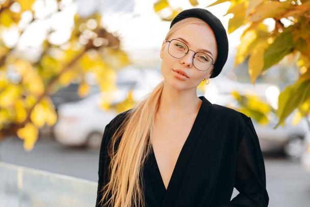 Portrait of a flirtatious girl with light daytime makeup, clear glasses on her eyes and a black beret, wearing a black blouse against a background of yellow-green leaves on a tree - Photo, Image
