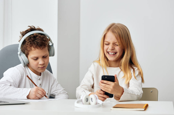 Szczęśliwe rodzeństwo odrabiające pracę domową online razem w domu. Chłopiec i dziewczynka siedzą przy stole w salonie, każdy z nich używa własnego laptopa i nosi słuchawki. - Zdjęcie, obraz