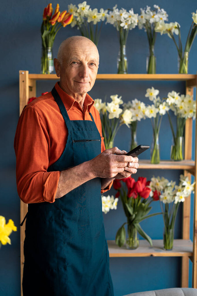 Ein älterer Mann, Blumenverkäufer, hält das Telefon mit einem Lächeln auf dem Gesicht. Antworten auf SMS von Kunden - Foto, Bild
