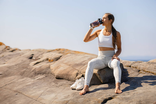 Encaixe a mulher nova no sportswear hidrata bebendo a água ao sentar-se em uma paisagem litoral rochosa. Capturando um momento de merecido descanso sob um céu limpo. - Foto, Imagem