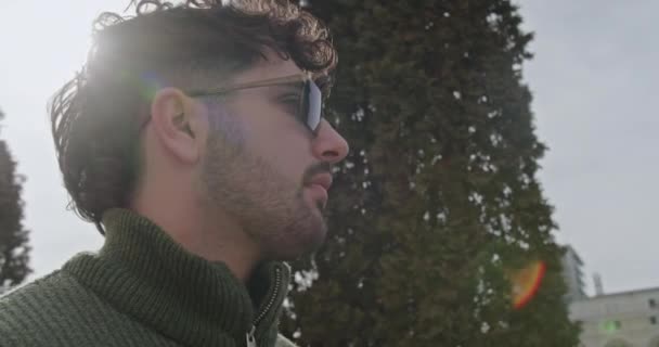 Een jonge man in een groene trui doet zijn bril voorzichtig uit in een rustige buitenomgeving, wat suggereert een moment van reflectie of een pauze in zijn drukke dag.. - Video