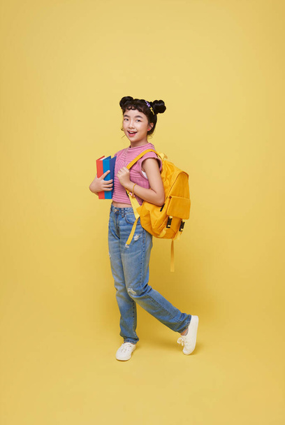 Портрет счастливой улыбчивой молодой азиатской ученицы начальной школы с блокнотом и рюкзаком готов к школе изолированы на желтом фоне. - Фото, изображение