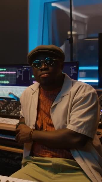 Medium vertical portrait of African American male music producer in hipster cap, sunglasses sitting in recording studio in front of professional digital audio workstation with editing software - Footage, Video