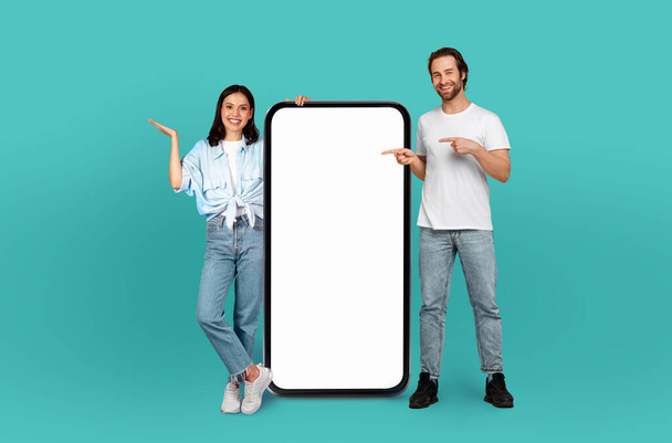 Een man en een vrouw staan zij aan zij en presenteren vrolijk een gigantische, lege smartphone-mockup, wat wijst op een promotie- of reclamecontext.. - Foto, afbeelding