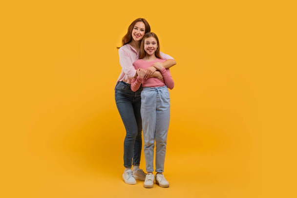 Mãe e filha estão de perto com um abraçando o outro por trás. Ambos estão sorrindo calorosamente e vestidos casualmente em jeans e tops leves contra um cenário amarelo vívido.. - Foto, Imagem