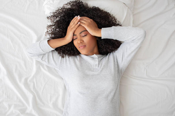 Die hispanische Frau liegt auf dem Rücken liegend im Bett, die Hände auf der Stirn in einer Geste, die auf Kopfschmerzen, Stress, Unwohlsein oder Erschöpfung hindeuten könnte - Foto, Bild