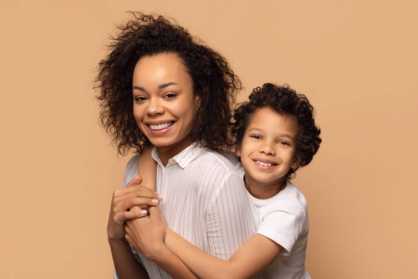 Uma mãe afro-americana alegre com cabelo encaracolado está segurando seu filho jovem amorosamente, pois ambos sorriem amplamente. Eles estão posando contra um fundo bege neutro, close-up - Foto, Imagem